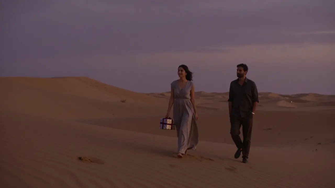 A woman walks alone across a desert dune at twilight, smiling gently as a man follows respectfully in the distance.